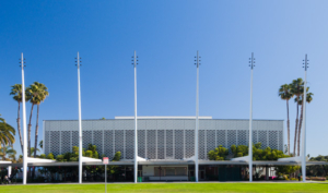 A large modernist public building faced with a curtain wall of windows and a cast-concrete grill, as well as five sky-piercing concrete masts, is framed by a blue sky and green lawn.