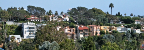  Two horizontal rows of large multi-story homes, including several from the early 20th century, are viewed from the opposite side of a canyon. Lush green landscaping and mature trees surround the architecture.