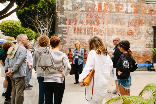 A Downtown walking tour docent faces talks to a small tour group while standing in front of an exterior brick wall covered with faded advertisements. 