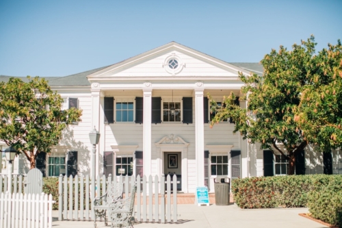 The south elevation of the Marion Davies Guest House, clad in white siding with black shutters and a grand two-story columned portico, on a sunny day.