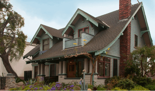  Double beams support a steeply pitched roof punctuated by two gables over the porch of a modest two-story craftsman home. The exterior is finished with white clapboard siding with green trim. 