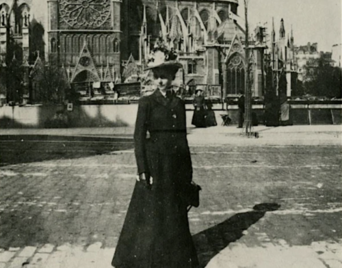 Black and white photo of architect Julia Morgan in a black dress and hat, standing in front of Notre Dame Cathedral.