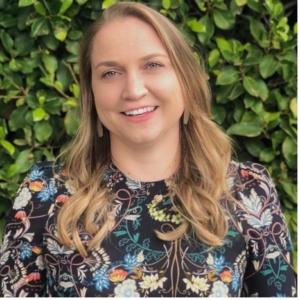 With a slight tilt to her smiling face, Catherine Conkle poses wearing a floral-print shirt in front of a thick green hedge. 