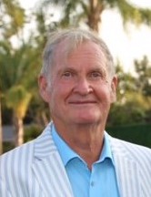 Local historian Mark Gorman, wearing a light blue shirt and gray-striped blazer, smiles against a background of palm trees and hedge work, his silver hair slightly tousled on his forehead. 