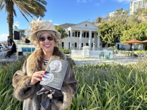 Wearing sunglasses and a wide smile, with a patch of native plants behind her, Robin Venturelli poses in a flapper-era fur coat and frilly beige hat while holding a biography of Marion Davies, the silent screen star Robin portrays annually at the Marion Davies Guest House, seen in the background. 