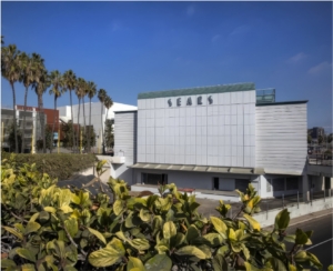 The clean Moderne lines of Santa Monica’s Sears Building gleam in the afternoon sun, embraced by a row of palms on its left and a hedge of tropical greenery in the foreground. 